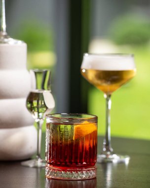 Portakal kabuğu garnitürlü klasik bir kırmızı kokteyl koyu renkli bir bar yüzeyindeki diğer sofistike içkilerin arasında yer alan desenli bir kaya bardağına yerleştirilir..