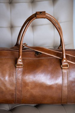 Eski tarz kahverengi deri bir spor çantası krem renkli bir sandalyenin üzerinde bulanık ahşap ve gri bir arka plana dayanır..