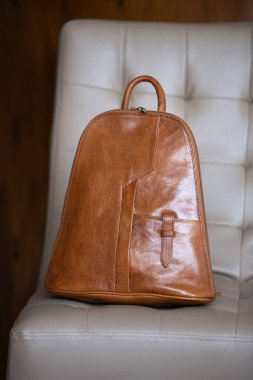 Açık krem rengi bir sandalyede kahverengi bir arka planda ince detaylarıyla bronzlaşmış deri bir sırt çantası duruyor.