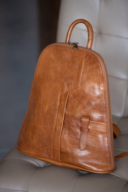 Açık krem rengi bir sandalyede kahverengi bir arka planda ince detaylarıyla bronzlaşmış deri bir sırt çantası duruyor.