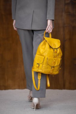 Gri bir takım elbiseyle poz veren şık, genç bir kadın, sıcak ahşap bir arka plana karşı parlak sarı bir sırt çantası tutuyor..