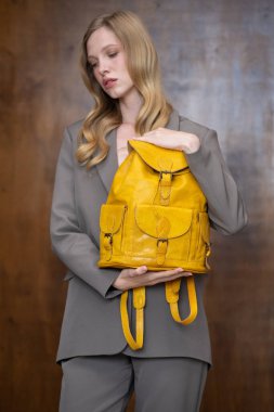 Uzun sarı saçlı şık bir genç kadın, gri bir takım elbise içinde, sıcak ahşap bir arka plana karşı canlı sarı bir deri çanta tutuyor..