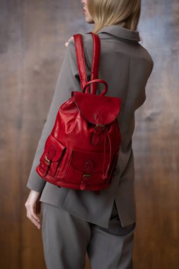 Uzun sarı saçlı şık bir genç kadın, gri bir takım elbise içinde, sıcak ahşap bir arka plana karşı canlı kırmızı bir deri çanta tutuyor..