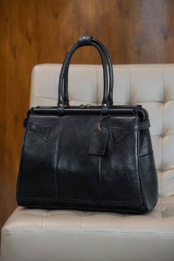 Antika bir siyah deri çanta, bulanık ahşap ve gri bir arka plana karşı krem renkli bir sandalyenin üzerinde duruyor..