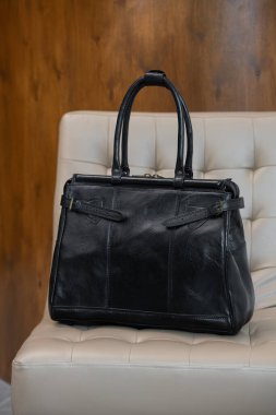 Antika bir siyah deri çanta, bulanık ahşap ve gri bir arka plana karşı krem renkli bir sandalyenin üzerinde duruyor..