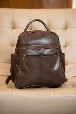 İnce detaylı kahverengi deri bir sırt çantası açık kremalı sandalyenin üzerinde duruyor.