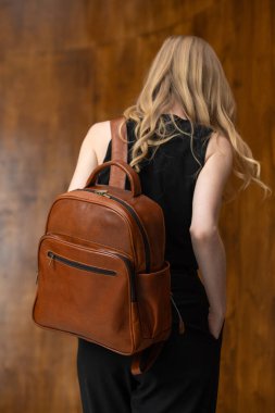 Uzun dalgalı sarı saçlı şık bir kadın model, sırt üstü bir perspektiften çekiliyor, omzunun üstünden bakıyor, ve kendine güvenen kahverengi bir sırt çantası modelliyor.