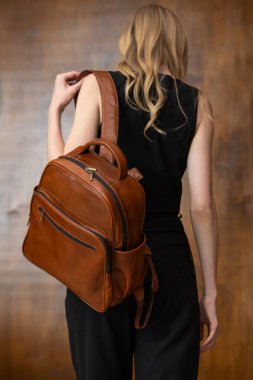 Uzun dalgalı sarı saçlı şık bir kadın model, sırt üstü bir perspektiften çekiliyor, omzunun üstünden bakıyor, ve kendine güvenen kahverengi bir sırt çantası modelliyor.