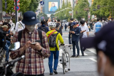 Tokyo, Japonya, 5 Kasım 2023: Sarı ceketli bisikletçi kalabalık bir caddede bisiklet taşıyor