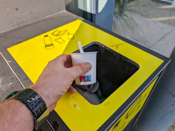 France, 10 August 2024: Hand throwing paper cup into yellow recycling bin