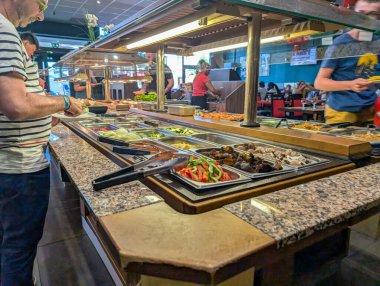 France, 03 August 2024 : Buffet patrons serving themselves food at a busy restaurant salad bar clipart