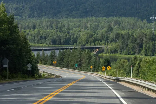Quebec, Kanada, 20 Temmuz 2025: Kavisli yol uzak tepelere doğru ormanlık nehir köprüsünden geçiyor