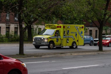 Quebec, Kanada, 07 Temmuz 2025: Ağaçlar ve arabalarla çevrili şehir caddesinde yürüyen sarı ambulans.