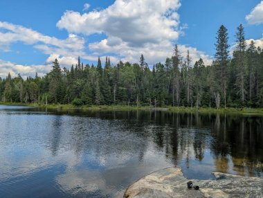 Quebec, Kanada, 18 Temmuz 2025: Huzurlu göl kenarındaki kayalık kıyıda orman yansımalı kamera