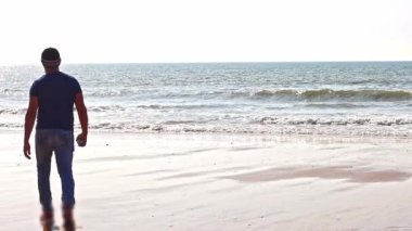 Mavi tişörtlü, kot pantolonlu ve şapkalı olgun bir adam sırtı kameraya dönük deniz kıyısında yürüyor. Arka planda, dalgalı deniz yavaş çekimde çekiliyor. Yalnızlık hissi veriyor..
