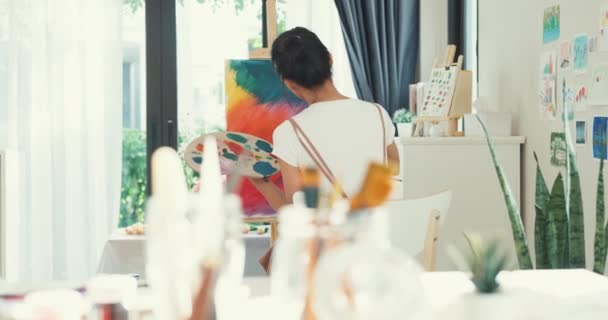 快乐的年轻亚洲女性穿着围裙 手握调色板 在家里舒适的车间里用画布在画布上创作艺术品 当代画家摘要现代艺术 创造力与人的概念 — 图库视频影像