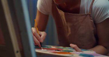 Genç Asyalı kadın palet tutarken önlük takıyor tuvale fırça boyası sürüyor geceleri evde rahat bir atölye çalışması yapıyor. Çağdaş Ressam Soyut Modern Sanat, Yaratıcılık ve İnsan Konsepti.