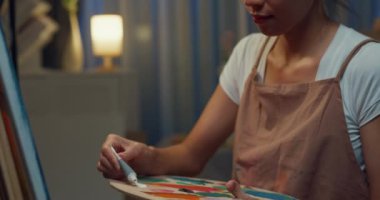 Genç Asyalı kadın palet tutarken önlük takıyor tuvale fırça boyası sürüyor geceleri evde rahat bir atölye çalışması yapıyor. Çağdaş Ressam Soyut Modern Sanat, Yaratıcılık ve İnsan Konsepti.