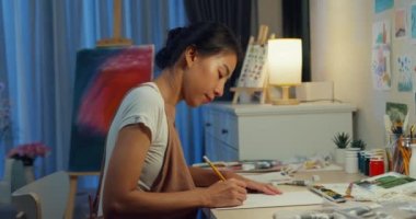 Genç Asyalı kadın önlük takıyor çizim defterine resim çiziyor geceleri evde rahat bir atölye çalışması yapıyor. Çağdaş Ressam Soyut Modern Sanat, Yaratıcılık ve İnsan Konsepti.