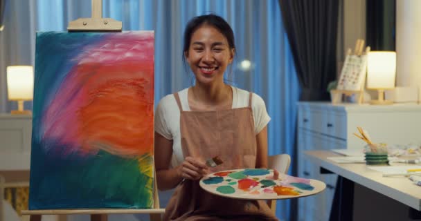 年轻快乐的亚洲女性穿着围裙微笑着看着相机 手握调色板站在帆布后面 晚上在家里画着舒适的艺术品作坊 当代画家抽象艺术 创意概念 — 图库视频影像
