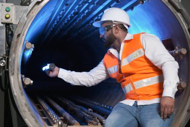 Güney Asyalı erkek fabrika işçisi veya insan mühendisi veya uzman personel güvenlik yeleği ve bakım kaskı giyer ve UV temizleme ile buhar kazanını kontrol ederler.