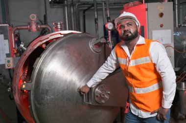 Güney Asyalı erkek fabrika işçisi veya mühendis veya uzman personel güvenlik yeleği ve bakım kaskı giyer ve buhar kazanını kontrol ederler.