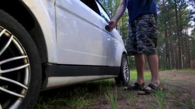 Bir adam küçük, beyaz bir arabanın kapısını açar ve içeri girer. Araba bir ormana park edilmiş..