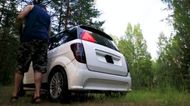 Bir adam ormanda park etmiş küçük beyaz bir arabaya yaklaşır ve bagajını açar..