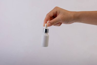 Elinde şeffaf bir arka planda duran beyaz bir cam damlası şişesi var. 