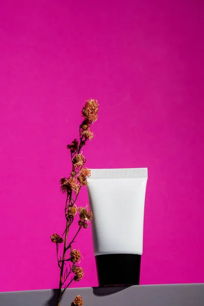 Plastic white tube for cream or lotion. Skin care or sunscreen cosmetic and dried flowers on pink background