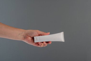 Krem veya losyon için plastik beyaz tüp. Gri arka planda eli olan cilt bakımı veya güneş kremi kozmetiği.