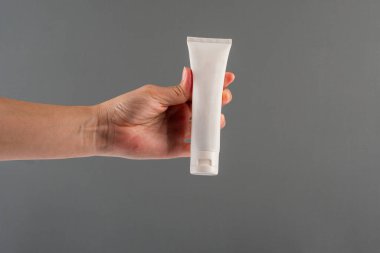 Krem veya losyon için plastik beyaz tüp. Gri arka planda eli olan cilt bakımı veya güneş kremi kozmetiği.