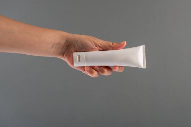 Krem veya losyon için plastik beyaz tüp. Gri arka planda eli olan cilt bakımı veya güneş kremi kozmetiği.