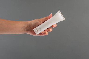 Krem veya losyon için plastik beyaz tüp. Gri arka planda eli olan cilt bakımı veya güneş kremi kozmetiği.