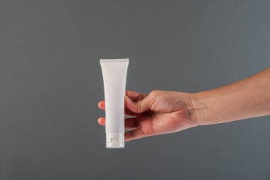 Krem veya losyon için plastik beyaz tüp. Gri arka planda eli olan cilt bakımı veya güneş kremi kozmetiği.