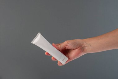 Krem veya losyon için plastik beyaz tüp. Gri arka planda eli olan cilt bakımı veya güneş kremi kozmetiği.