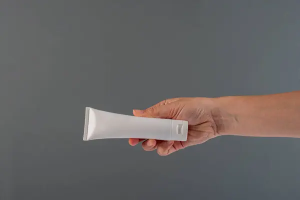 Krem veya losyon için plastik beyaz tüp. Gri arka planda eli olan cilt bakımı veya güneş kremi kozmetiği.