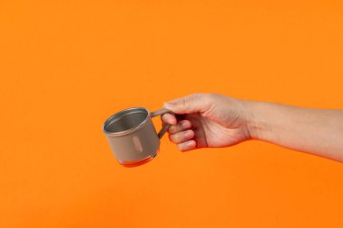 Eldeki paslanmaz çelik bardak turuncu arka planda izole edilmiş.