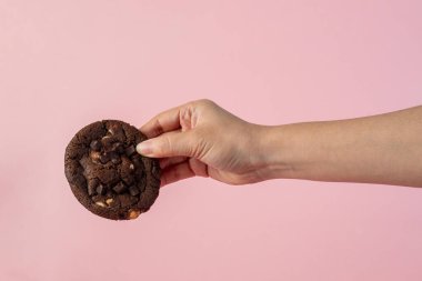 Üzerinde çikolata parçacıkları ve el olan büyük bir çikolatalı kurabiye, pembe arka planda..