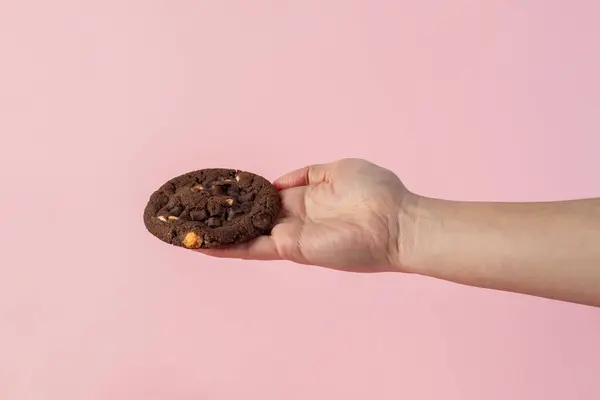 Üzerinde çikolata parçacıkları ve el olan büyük bir çikolatalı kurabiye, pembe arka planda..