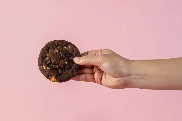 Üzerinde çikolata parçacıkları ve el olan büyük bir çikolatalı kurabiye, pembe arka planda..