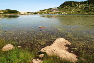 View over the Fish Lake, Ribnoto Ezero, water plants, grass can be seen below the smooth surface, the Seven Lakes Chalet can be seen on the shore in the background reflecting in the water, Rila National Park, close to Sofia, Bulgaria 2022 clipart
