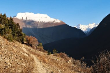 Yukarı Pisang köyünde küçük bir patika, Paungda Danda 'nın düz batı kayalık yüzü, Pisang' ın Büyük Duvarı ve arka planda Lamjung Kailas zirvesi, Annapurna Pisti, Nepal 2022