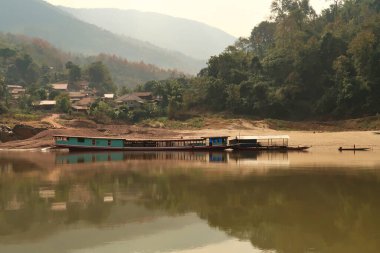 View from the slow boat from Thailand to Laos onto the shore of the Mekong River, another docked slow boat is reflecting in the smooth water in front of a local village, Laos 2023 clipart