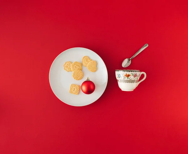Minimal composition with Christmas cookies and red bauble on white plate and tea cup with spoon. New year concept.