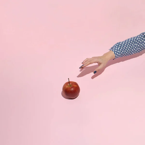 Female hand reaching for fresh apple on pink background. Minimal concept.