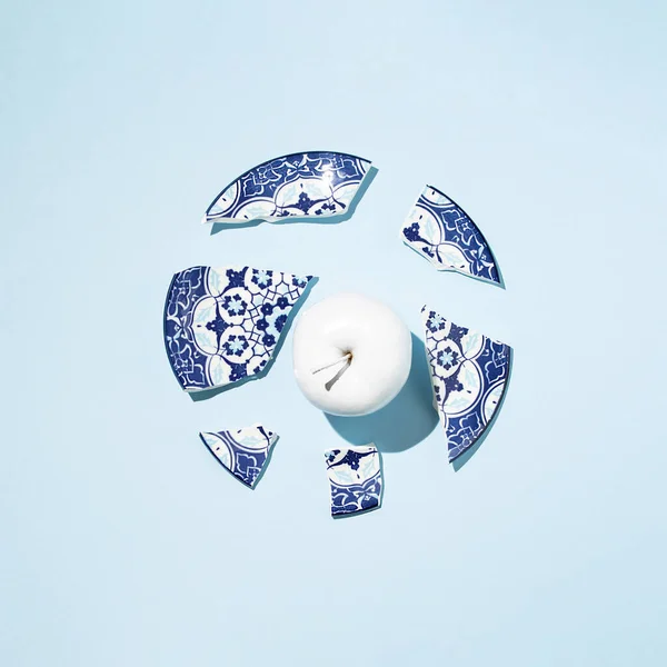 Fresh apple coloured in white paint and broken china porcelain plate on blue background.