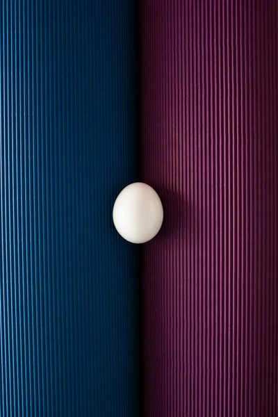 White colored egg between folded sheets of blue and purple corrugated cardboards. Minimal Easter concept.
