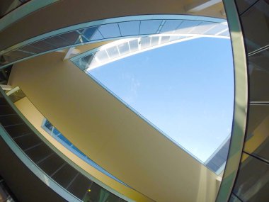 Karmaşık şekilli ve yaya geçitli modern bir mimari binanın altından görüntü. Yansıtıcı pencereler ve arka planda açık mavi gökyüzü. Şirket sahnelerini ya da mimari projelerin sunumunu temsil etmek için ideal resim