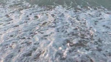 Yumuşak dalgaların ritmik hareketi kumlu bir sahilde kıyıya vuruyor. Köpüklü beyaz ibikler, pürüzsüz, ıslak kumla güzel bir tezat oluşturuyor, dingin ve zamansız bir sahne yaratıyor..